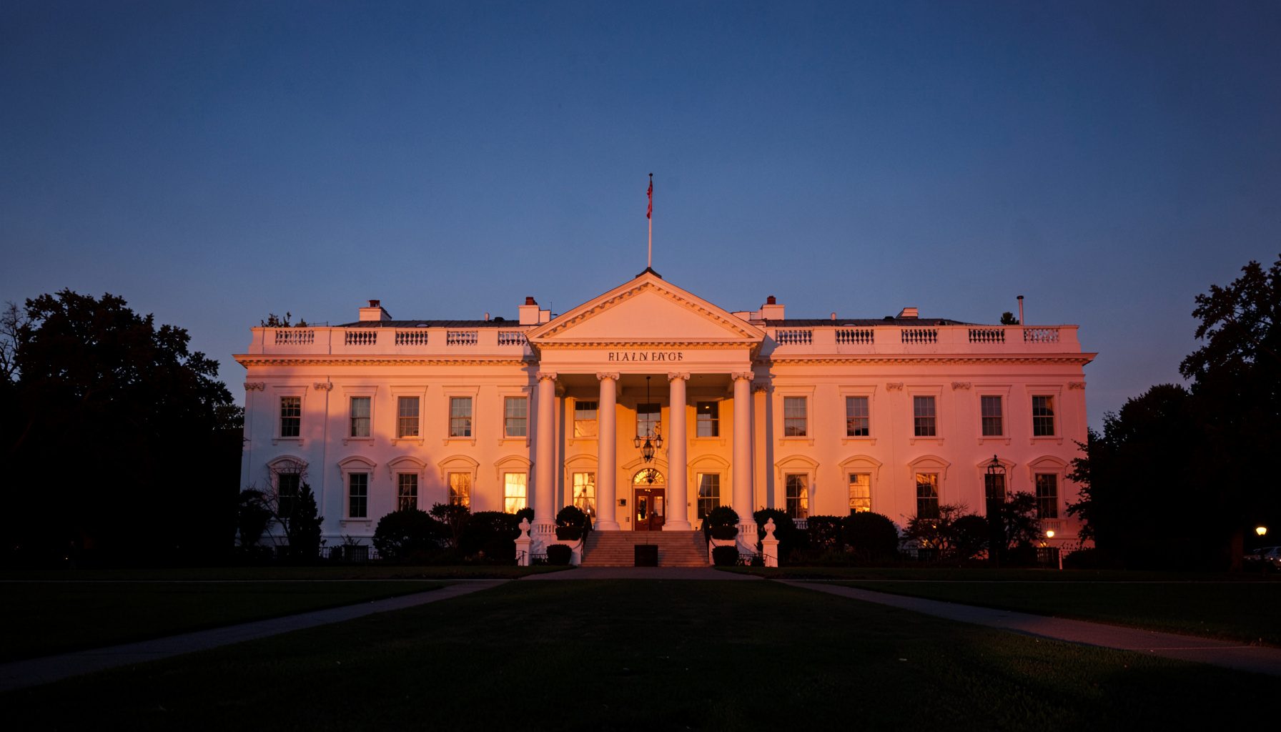 A photograph of the White House seen from across the South Lawn at dusk, with the building facade glowing warmly against a deepening blue sky.