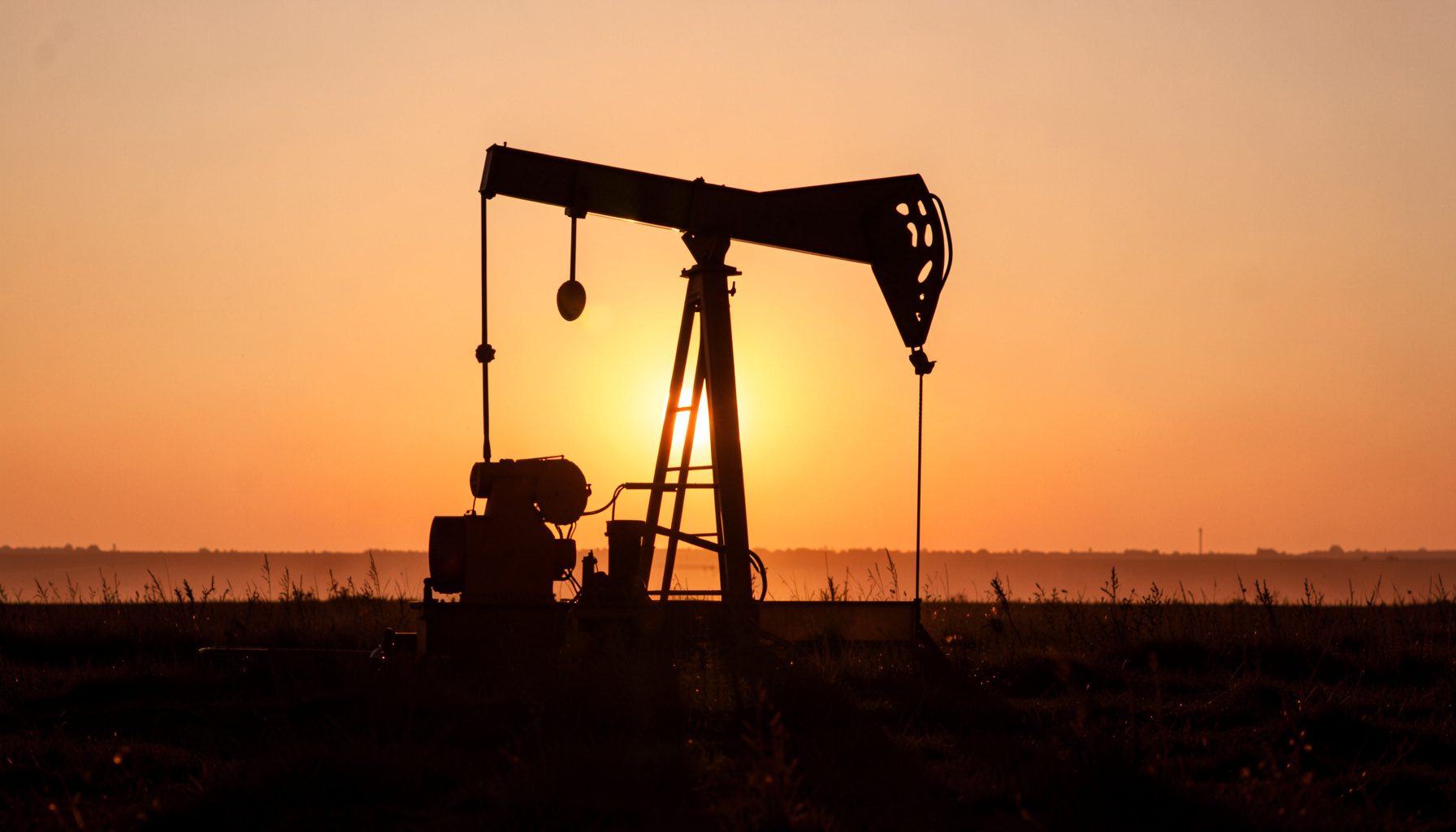 A photograph of an oil pump jack silhouetted against a golden sunset sky, with dry grassland stretching toward the horizon.