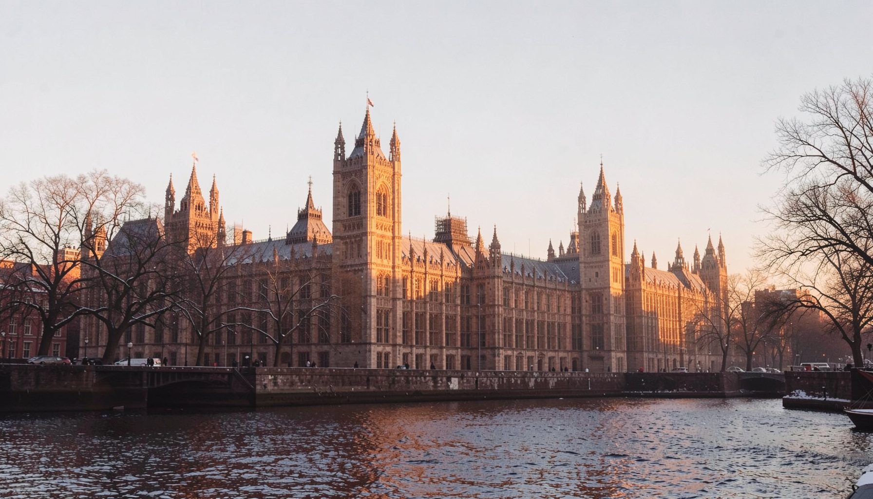 A photograph of the Houses of Parliament in London under a pale winter sky, with bare trees lining the Thames embankment in soft morning light.