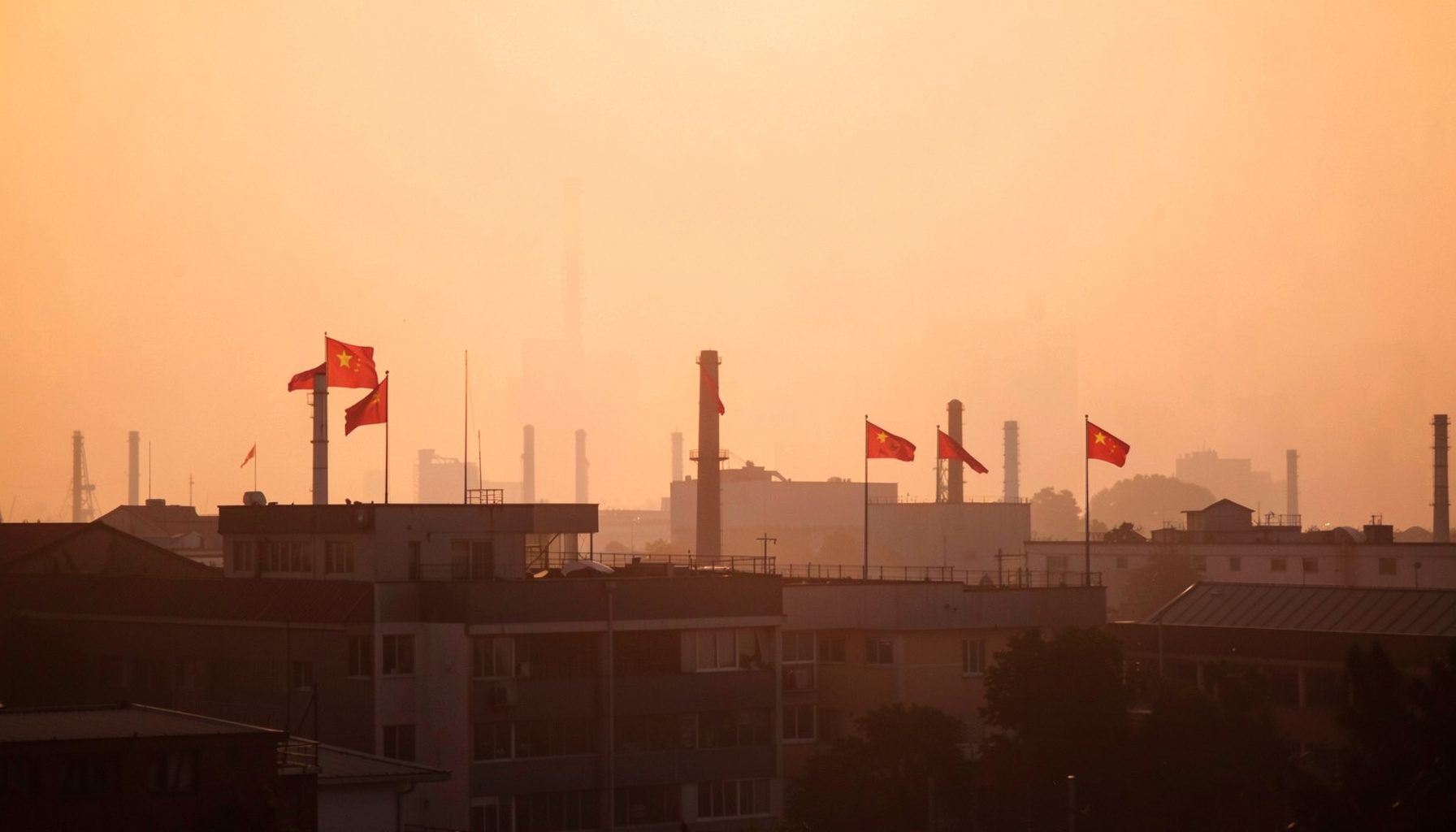 A photograph of factory buildings with Chinese flags visible, set against a hazy industrial skyline in the early morning light.